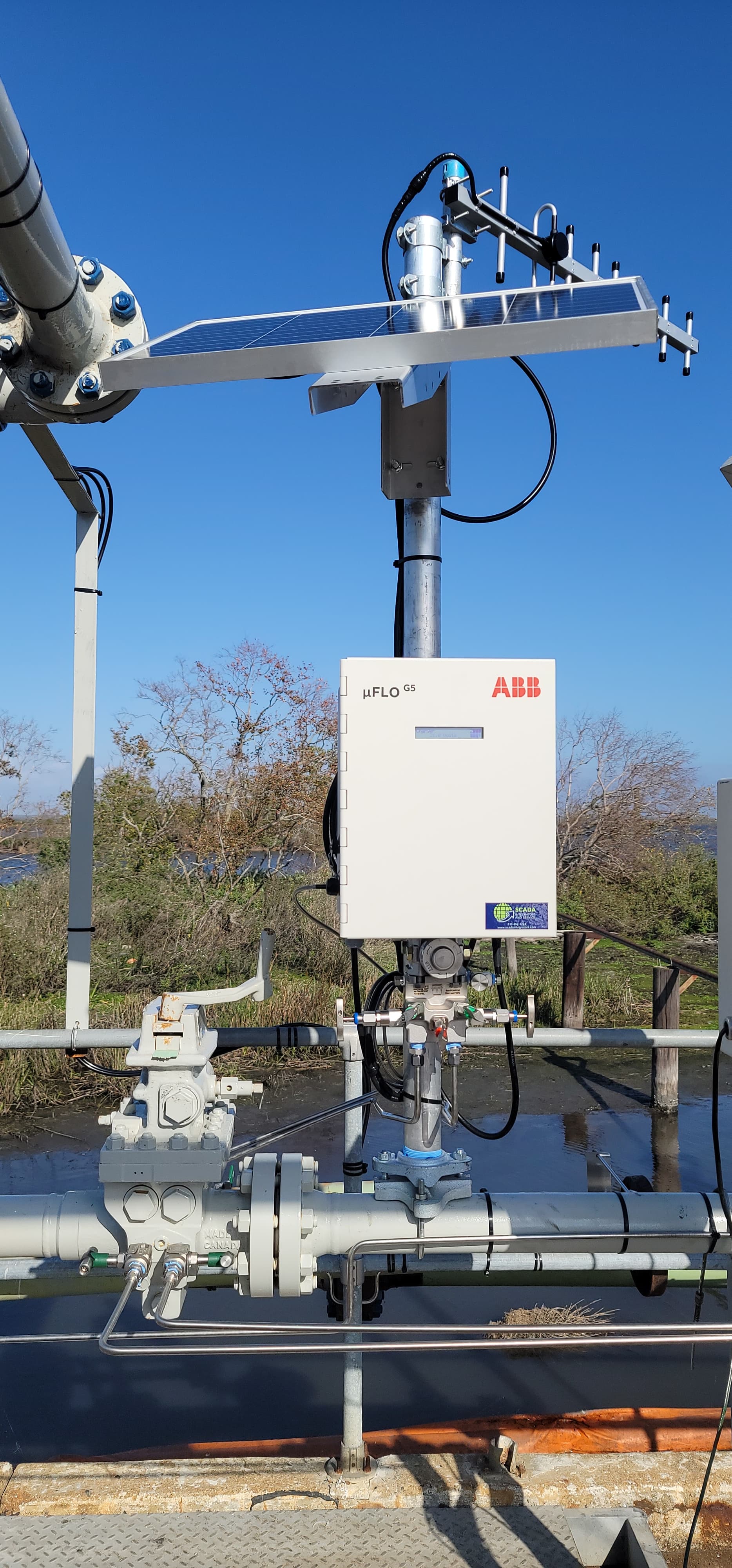 Solar-powered ABB flow computer and antenna mounted on industrial pipes in a sunny wetland.